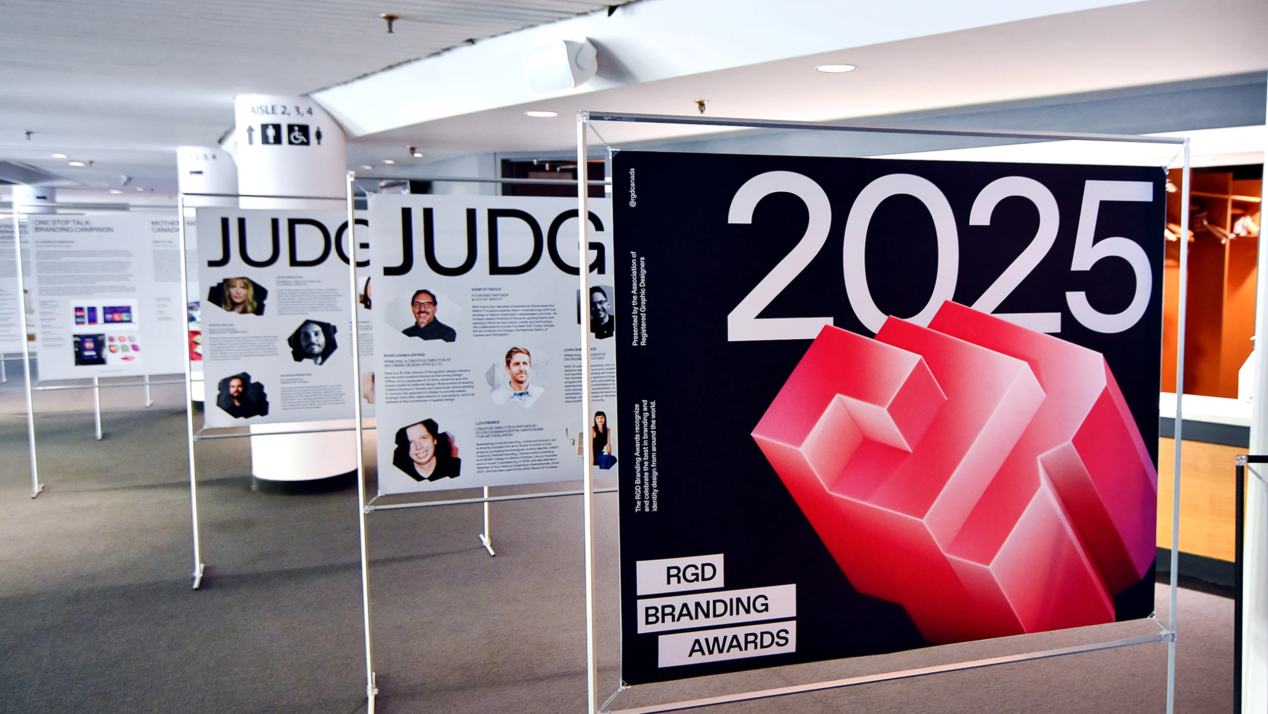 Environmental signage at a conference, displaying judge profiles and 2025 branding posters on metal stands.