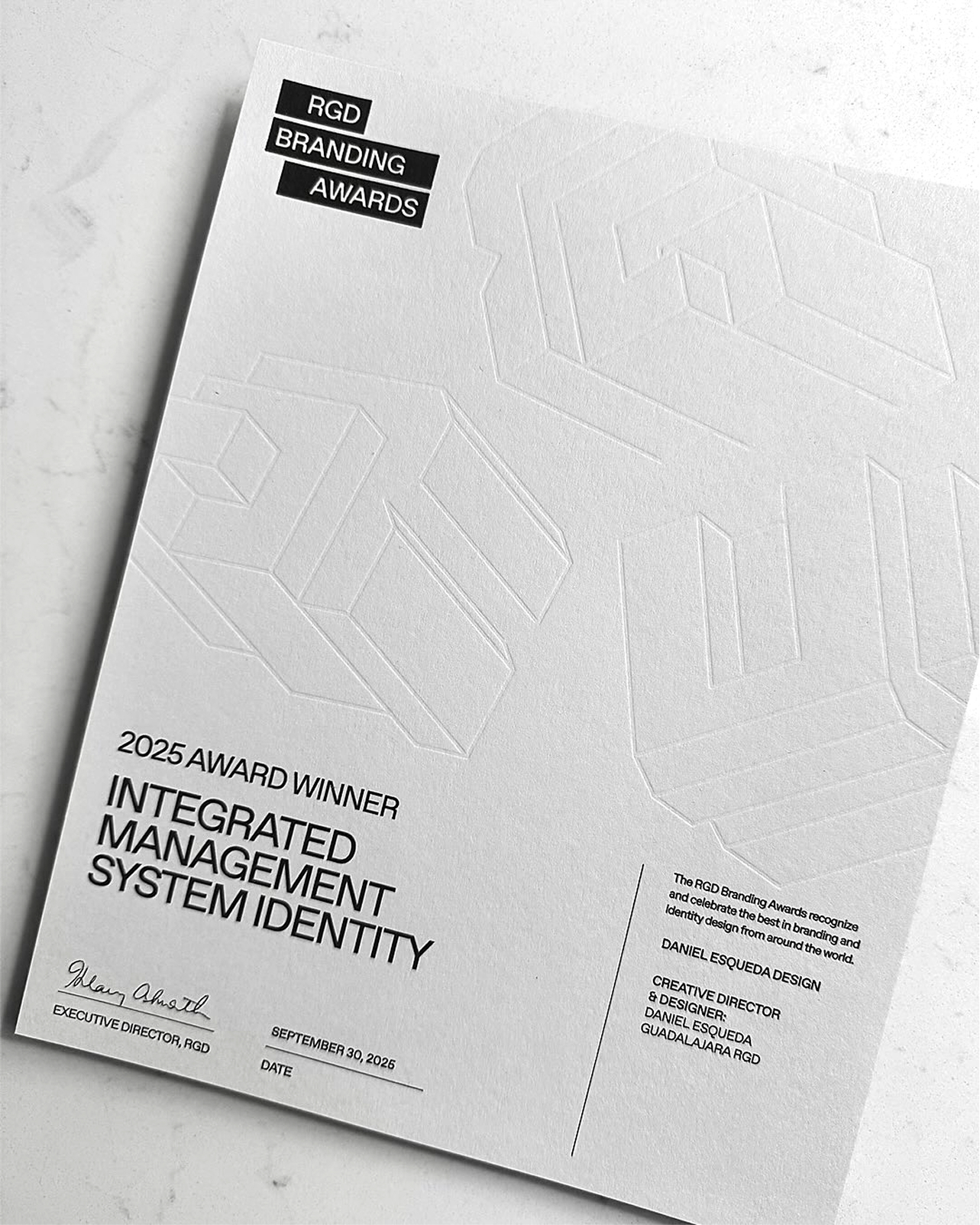 Close-up of white award certificate folder featuring blind embossed 3D geometric patterns and Favorit Regular typography.