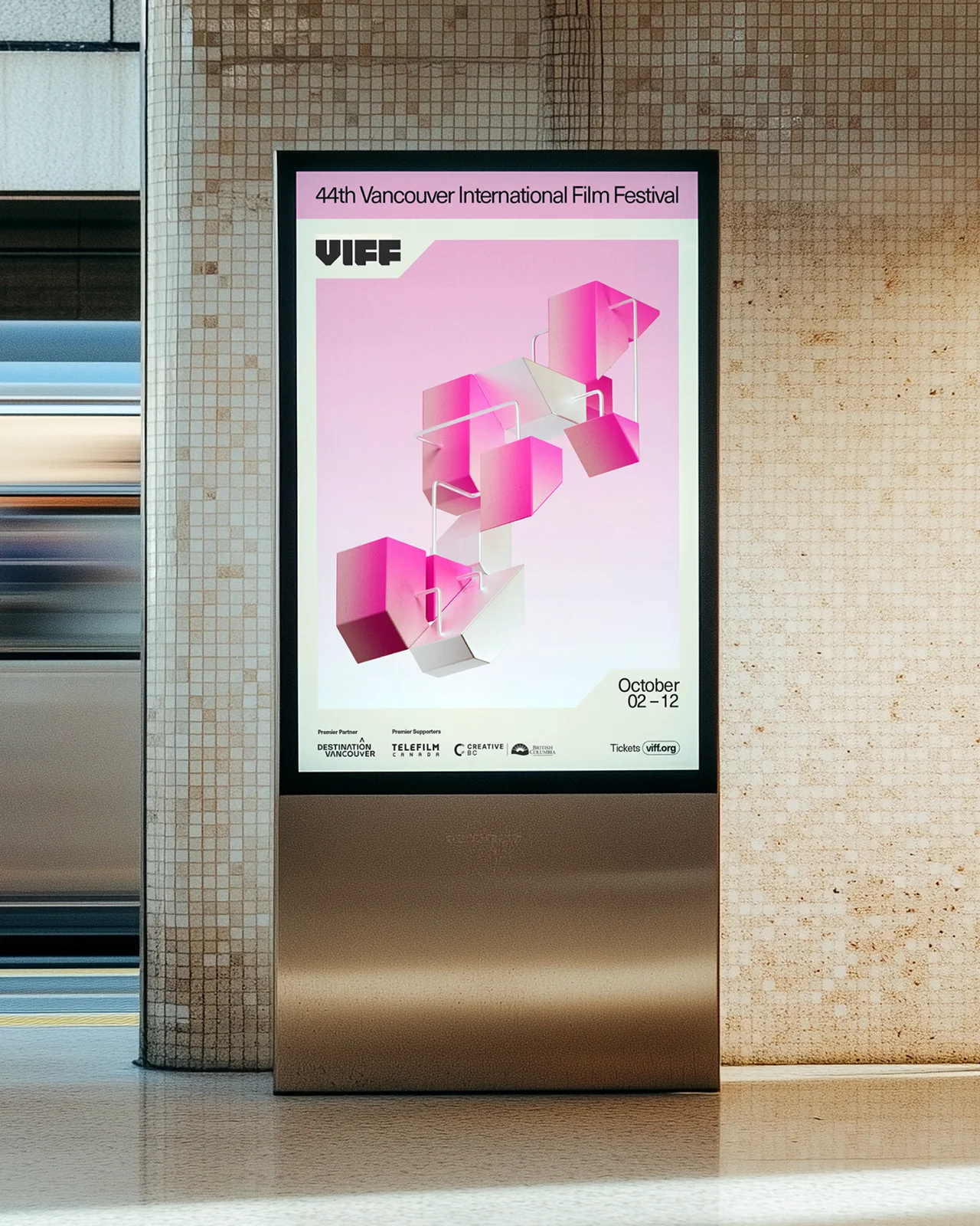 A vertical digital kiosk in a transit station showing a pink 3D modular brand graphic for VIFF 2025.