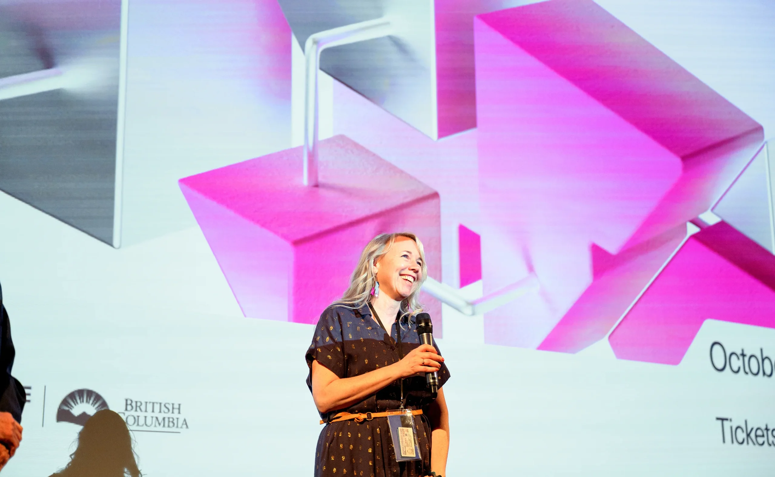 A presenter speaking in front of a massive cinema screen displaying pink 3D VIFF 2025 branding.