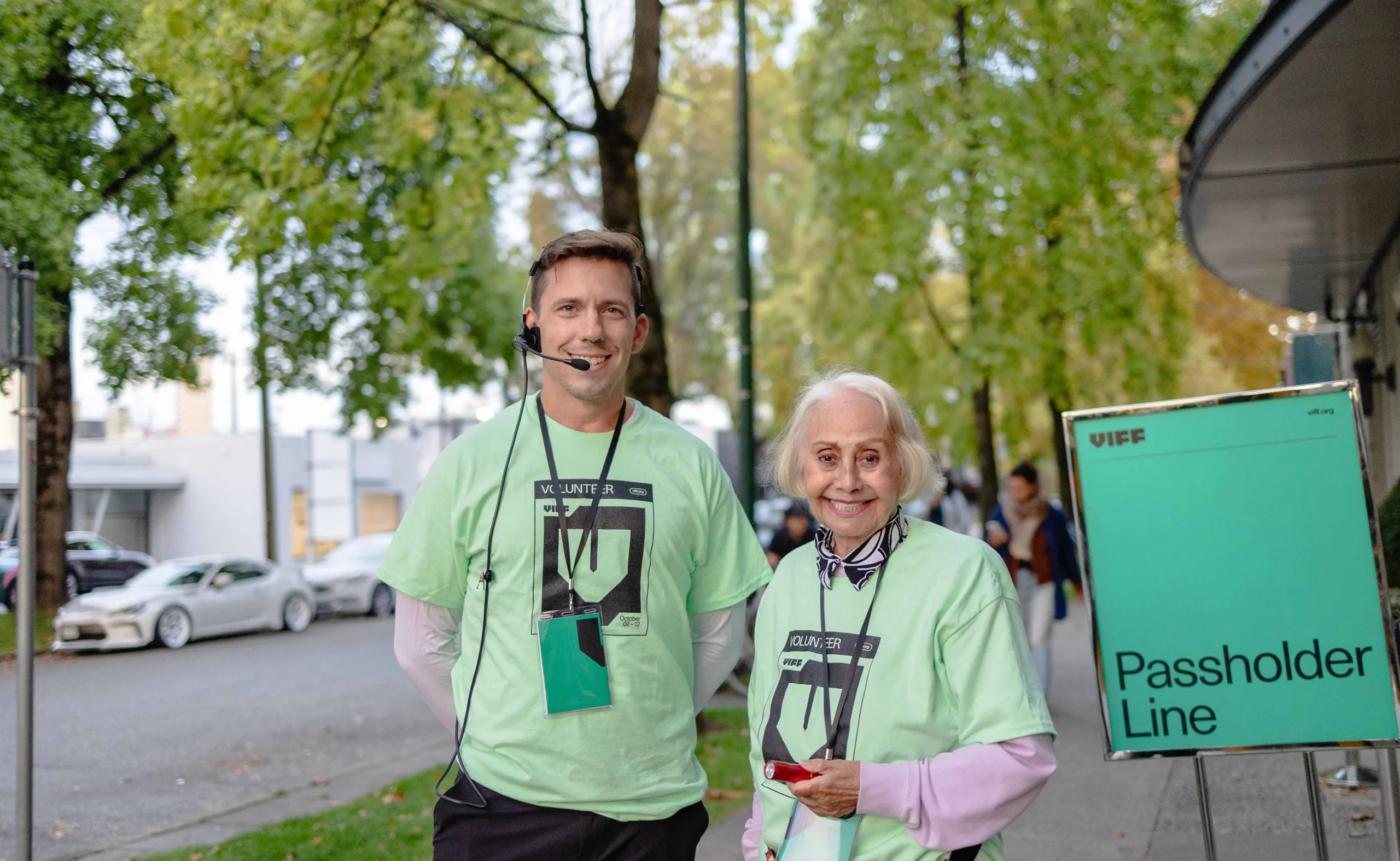 Two festival volunteers wearing light green VIFF 2025 branded t-shirts standing by a green Passholder Line sign.
