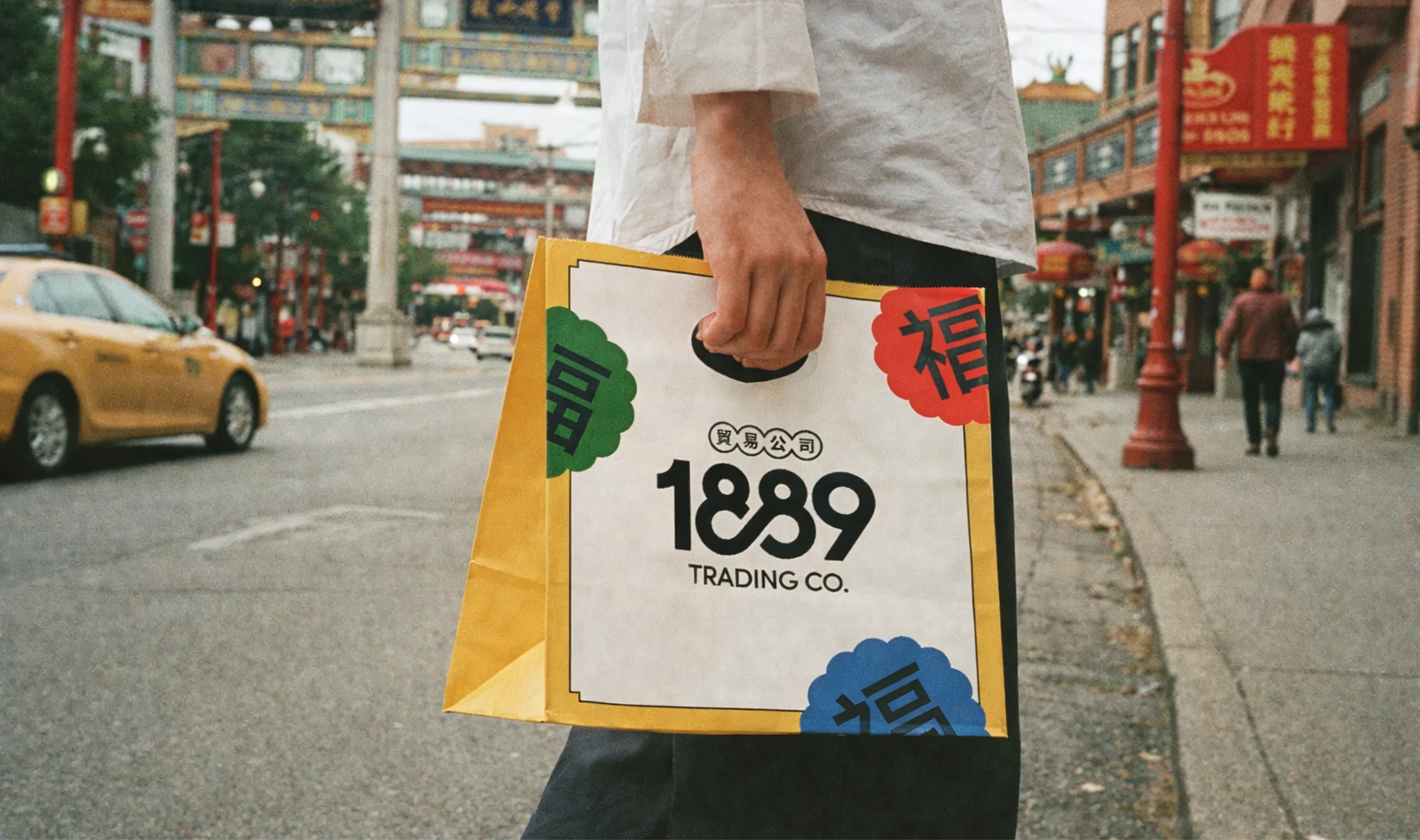 Person carrying a branded 1889 Trading Co. paper bag in front of the Chinatown Millennium Gate in Vancouver.