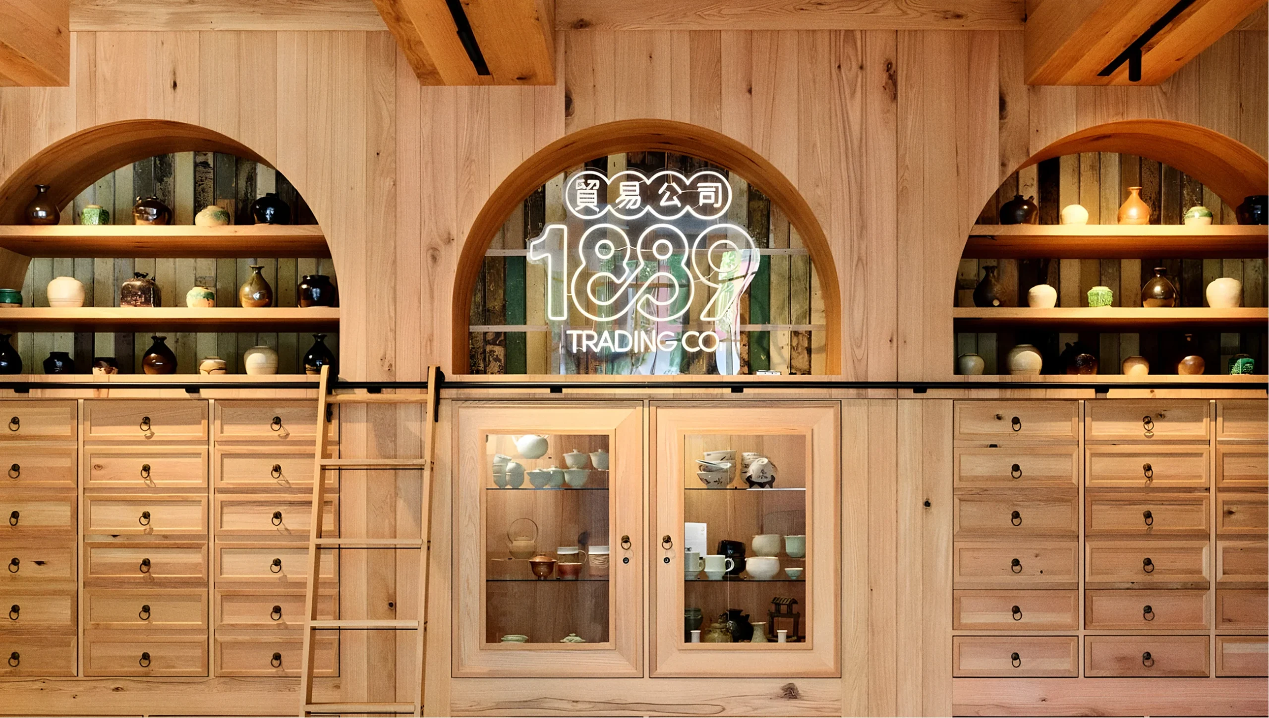 Modern retail interior with light wood cabinetry, apothecary drawers, and an arched neon 1889 Trading Co. logo.